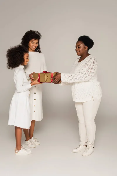 Tiro de longitud completa de la abuela afroamericana sonriente presentando caja de regalo a hija y nieta sobre fondo gris - foto de stock