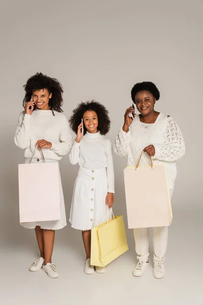 Aufnahme in voller Länge von afrikanisch-amerikanischer Mutter, Tochter und Großmutter, die Einkaufstüten in der Hand halten und auf grauen Hintergrund mit Mobiltelefonen sprechen — Stock Photo