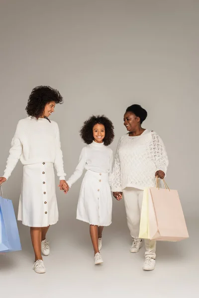 Lächelnde afrikanisch-amerikanische Mutter, Tochter und Großmutter gehen mit Einkaufstüten und halten Händchen auf grauem Hintergrund — Stockfoto