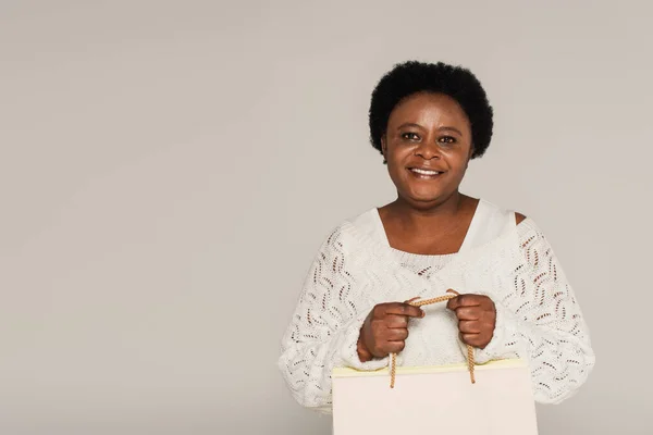 Souriant afro-américaine d'âge moyen en vêtements blancs tenant des sacs à provisions isolés sur gris — Photo de stock