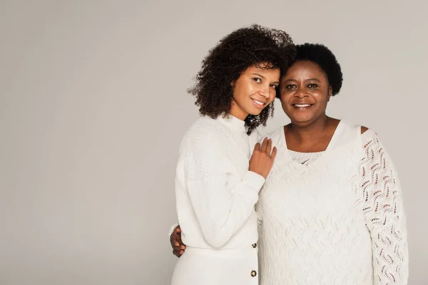 Hija adulta afroamericana sonriente abrazando cuidadosamente a la madre de mediana edad aislada en gris - foto de stock