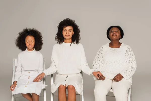 Афроамериканська дочка, мати і бабуся тримаються за руки, сидячи з закритими очима і молячись ізольовано на сірому — стокове фото