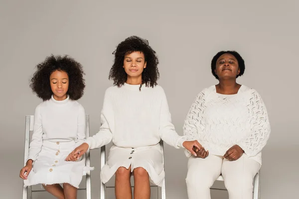 Hija afroamericana, madre y abuela cogidas de la mano, sentadas con los ojos cerrados y rezando aisladas en gris - foto de stock