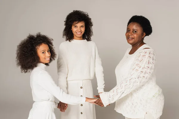 Fröhliche afrikanisch-amerikanische Tochter, Mutter und Großmutter halten Händchen und schauen in die Kamera auf grauem Hintergrund — Stockfoto