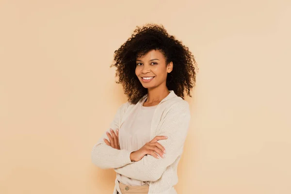 Sonriente mujer afroamericana de pie con los brazos cruzados aislados en beige - foto de stock
