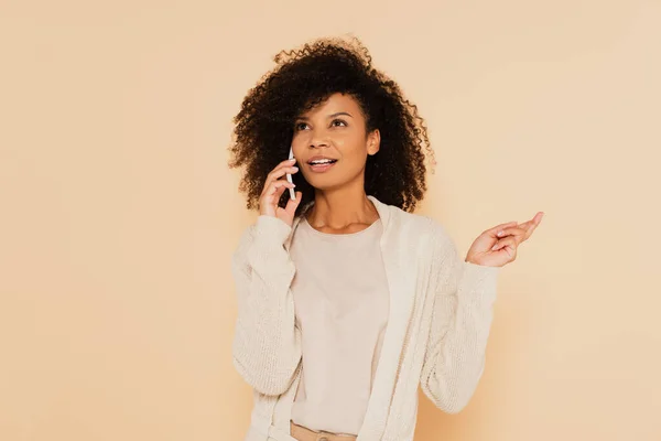 Thoughtful african american woman speaking on cellphone and gesturing with hand isolated on beige — Stock Photo