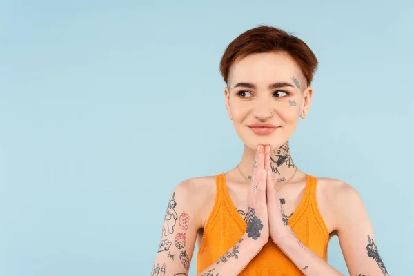 Задоволена і татуйована жінка з молитовними руками дивиться ізольовано на синьому — стокове фото
