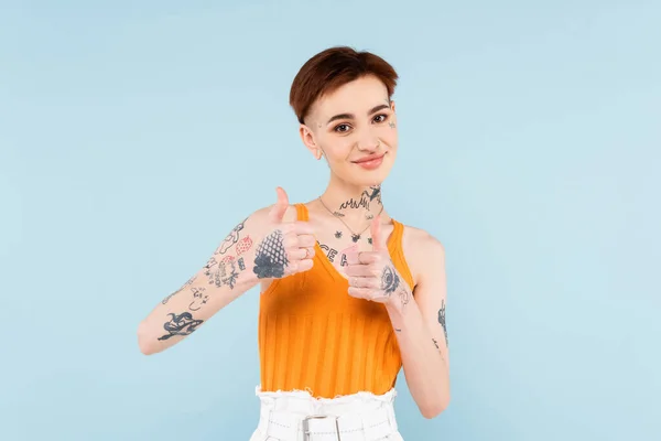 Pleased and tattooed woman showing thumbs up isolated on blue — Stock Photo