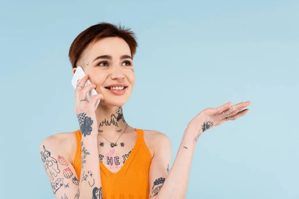 Весела і татуйована жінка говорить на смартфоні, жестикулюючи ізольовано на синьому — стокове фото
