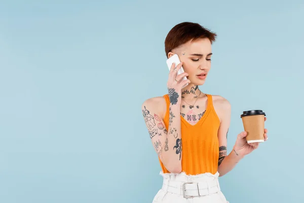 Tätowierte Frau spricht auf Smartphone, während sie Kaffee in der Hand hält, um sich auf blauem Grund zu isolieren — Stockfoto