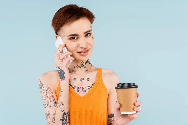 Mujer sonriente y tatuada hablando en el teléfono inteligente mientras sostiene el café para ir aislado en azul - foto de stock