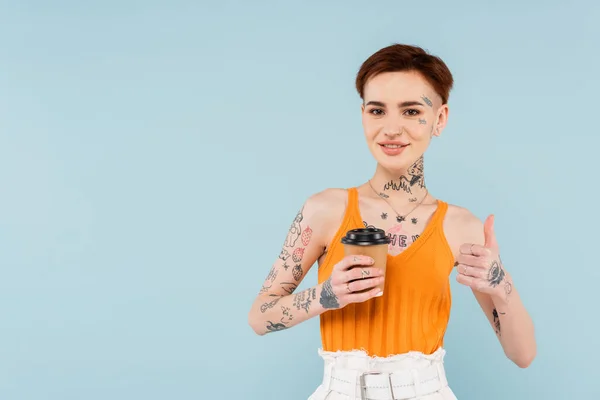 Mujer sonriente y tatuada mostrando el pulgar hacia arriba mientras sostiene la taza de papel aislada en azul - foto de stock