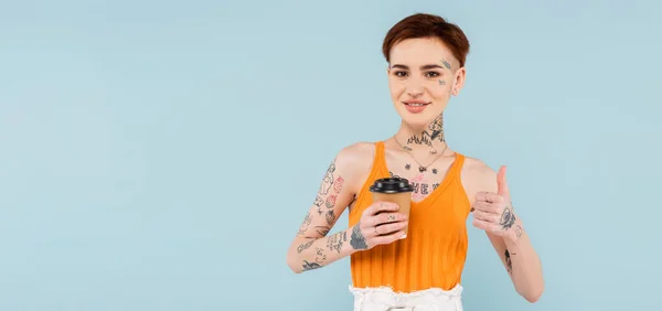 Mujer feliz y tatuada mostrando el pulgar hacia arriba mientras sostiene la taza de papel aislada en azul, pancarta - foto de stock