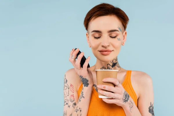 Mujer alegre y tatuada sosteniendo taza de papel y oliendo café para ir aislado en azul - foto de stock