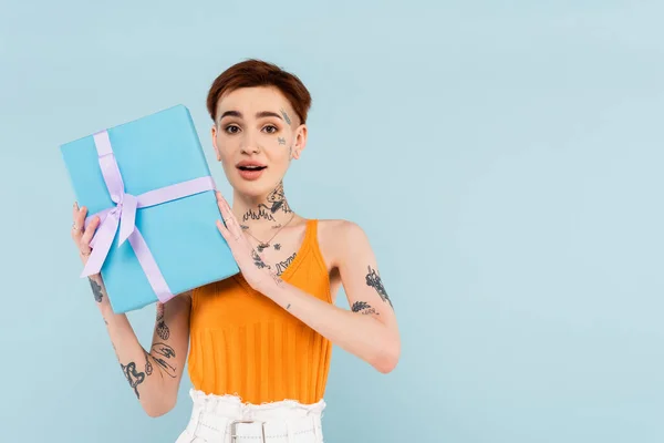 Здивована і татуйована жінка тримає загорнутий подарунок ізольовано на синьому — Stock Photo