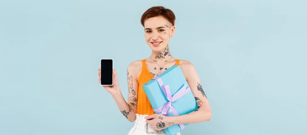Happy woman holding wrapped present and smartphone with blank screen isolated on blue, banner — Stock Photo