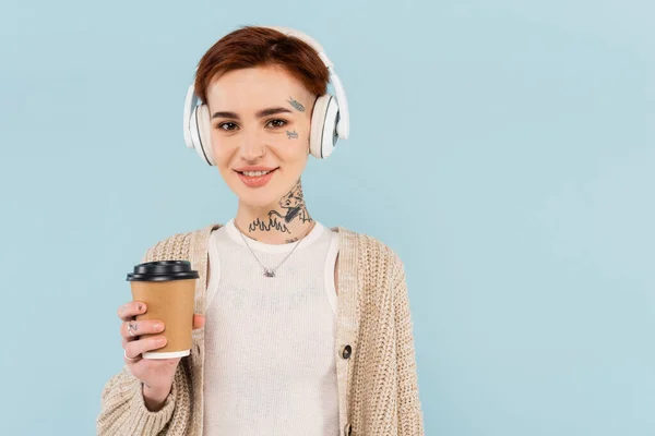 Усміхнена татуйована жінка в бездротових навушниках тримає паперову чашку ізольовано на синьому — стокове фото