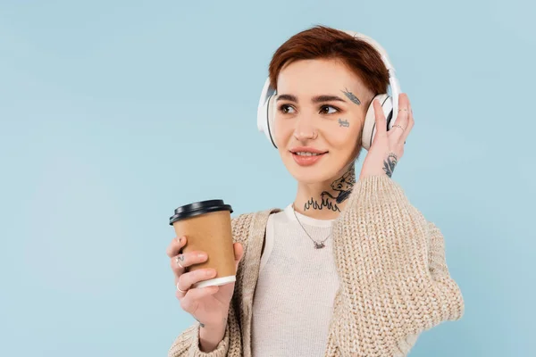Lächelnde Frau mit Tätowierungen in drahtlosen Kopfhörern, die Pappbecher isoliert auf Blau hält — Stockfoto