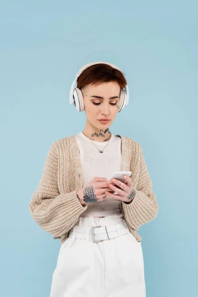 Mujer tatuada en auriculares inalámbricos charlando en un teléfono inteligente aislado en azul - foto de stock