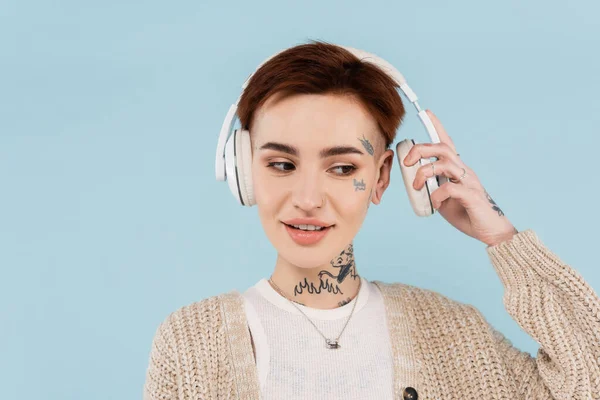 Усміхнена і татуйована жінка регулює бездротові навушники, дивлячись ізольовано на синьому — стокове фото