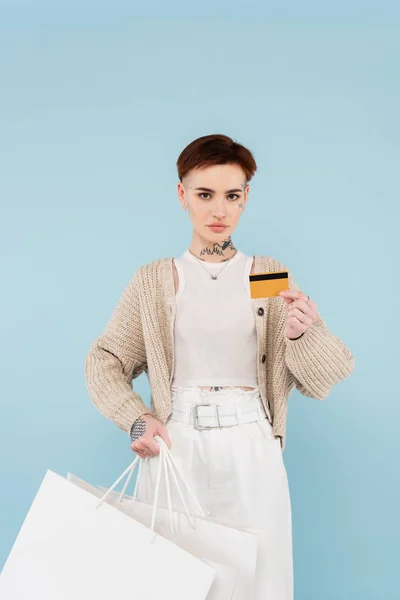 Joven mujer tatuada sosteniendo tarjetas de crédito y bolsas de compras aisladas en azul - foto de stock