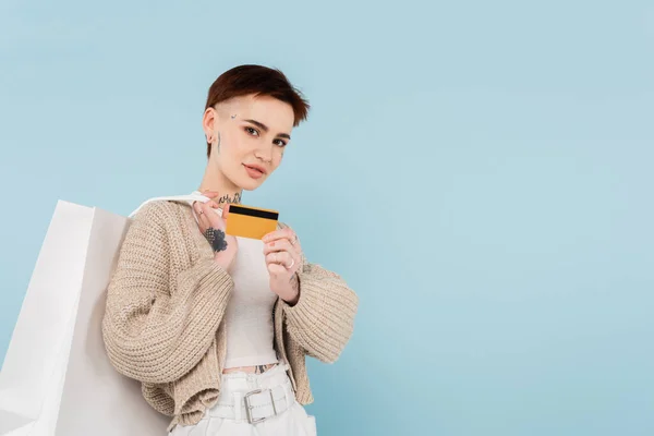 Young woman with tattoos holding credit card and shopping bags isolated on blue — Stock Photo