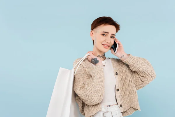 Feliz joven con tatuajes sosteniendo bolsas de compras mientras habla en un teléfono inteligente aislado en azul - foto de stock