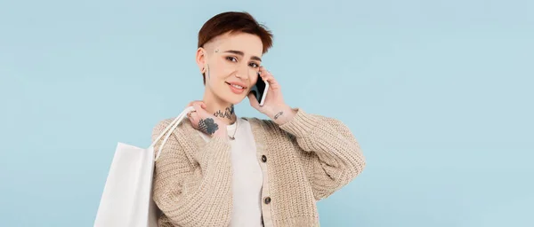 Feliz joven con tatuajes sosteniendo bolsas de compras mientras habla en un teléfono inteligente aislado en azul, pancarta - foto de stock