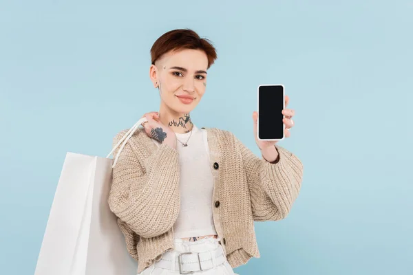Feliz joven con tatuajes sosteniendo bolsas de compras y teléfono inteligente con pantalla en blanco aislado en azul - foto de stock