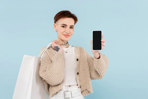 Jovem feliz com tatuagens segurando sacos de compras e smartphone com tela em branco isolado no azul — Fotografia de Stock