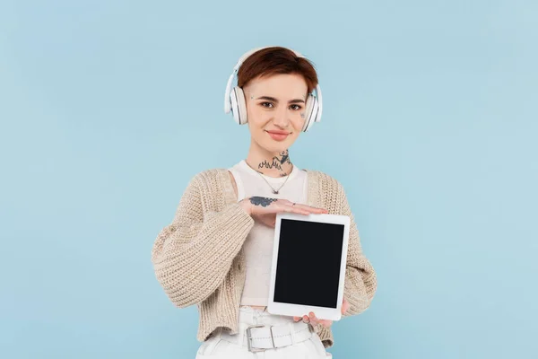 Happy woman with tattoos holding digital tablet with blank screen and listening music in wireless headphones isolated on blue — Stock Photo