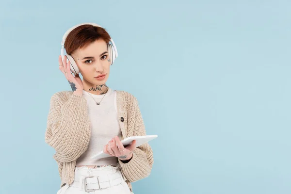 Junge Frau mit Tätowierungen, digitales Tablet in der Hand und Musik in drahtlosen Kopfhörern isoliert auf blauem Grund hörend — Stockfoto
