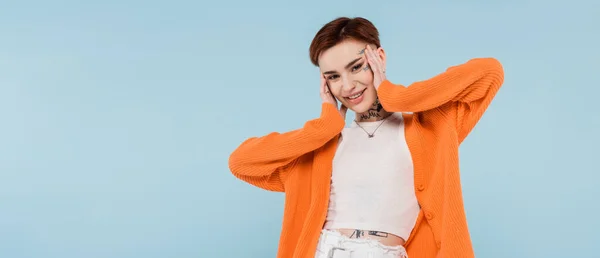 Feliz joven con tatuajes en cárdigan naranja posando aislado en azul, pancarta - foto de stock