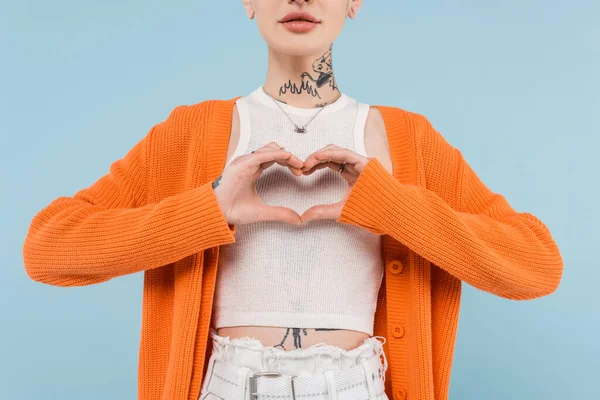 Vista ritagliata della giovane donna tatuata in cardigan arancione mostrando il cuore con le mani isolate sul blu — Foto stock