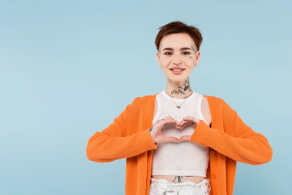 Happy young tattooed woman in orange cardigan showing heart with hands isolated on blue — Stock Photo