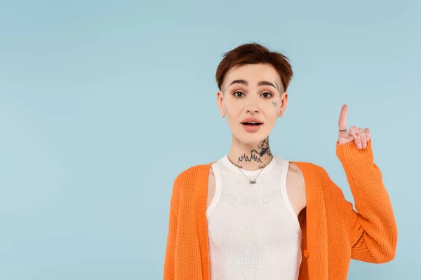 Amazed young tattooed woman in orange cardigan pointing up with finger isolated on blue — Stock Photo