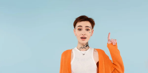 Amazed young tattooed woman in orange cardigan pointing up with finger isolated on blue, banner — Stock Photo