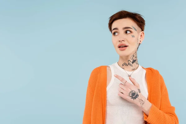 Amazed young tattooed woman in orange cardigan pointing away with finger isolated on blue — Stock Photo