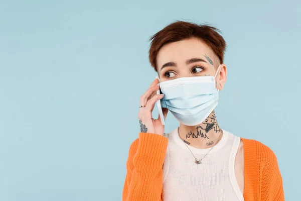 Mujer tatuada en cárdigan naranja y máscara médica hablando en teléfono inteligente aislado en azul - foto de stock