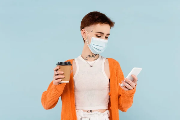 Joven mujer tatuada en máscara médica usando teléfono inteligente y sosteniendo taza de papel aislado en azul - foto de stock