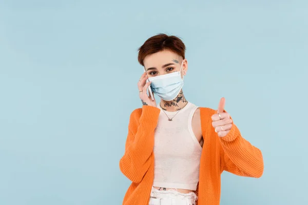 Young tattooed woman in orange cardigan and medical mask talking on smartphone while showing thumb up isolated on blue — Stock Photo