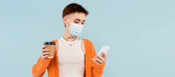 Giovane donna tatuata in maschera medica utilizzando smartphone e tenendo coppetta di carta isolata su blu, banner — Foto stock