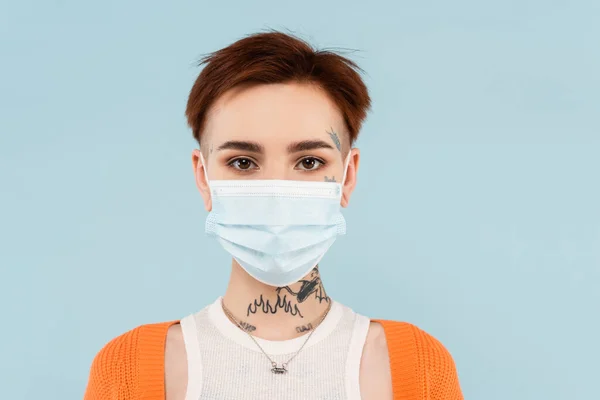 Mujer joven tatuada en cárdigan naranja y máscara médica aislada en azul - foto de stock