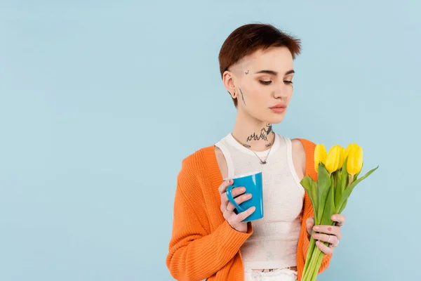 Young tattooed woman in orange cardigan holding cup of coffee and yellow tulips isolated on blue — Stock Photo