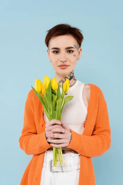 Young tattooed woman in orange cardigan holding yellow tulips isolated on blue — Stock Photo