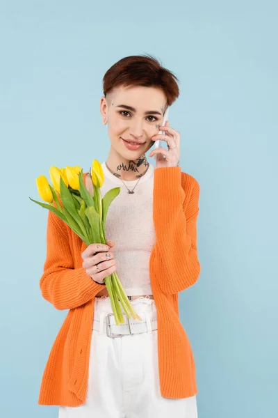 Молода татуйована жінка в помаранчевому кардигані тримає жовті тюльпани, розмовляючи на смартфоні ізольовано на синьому — стокове фото