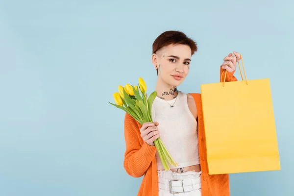 Молода татуйована жінка в помаранчевому кардигані тримає жовті тюльпани і сумку для покупок ізольовано на синьому — стокове фото