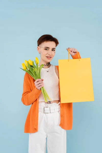 Femme heureuse avec des tatouages dans un cardigan orange tenant des tulipes jaunes et un sac à provisions isolé sur bleu — Photo de stock