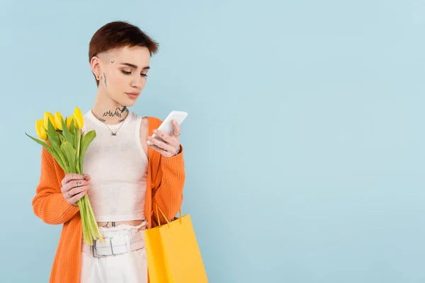 Donna con tatuaggi in possesso di tulipani gialli e shopping bag mentre chatta su smartphone isolato su blu — Foto stock