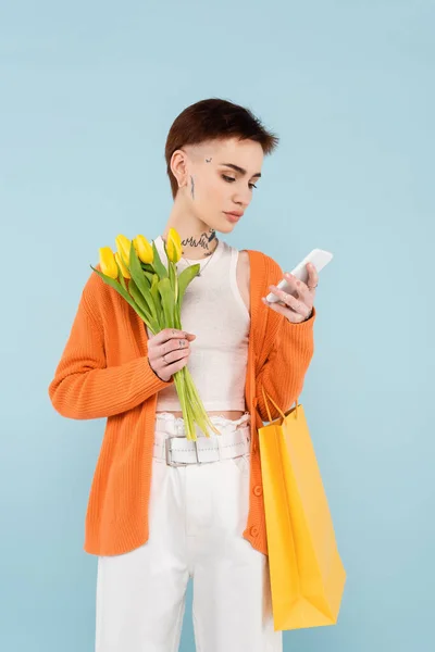 Mujer joven con tatuajes sosteniendo tulipanes amarillos y bolsa de compras mientras chatea en el teléfono celular aislado en azul - foto de stock